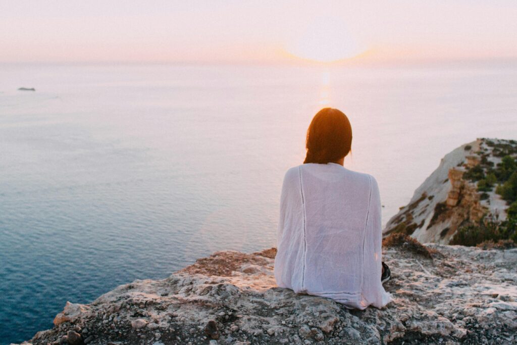 pexels-photo-185801-185801 A serene view of a woman sitting on a rocky cliff, gazing at the ocean during sunset. Perfect for relaxation and travel themes.