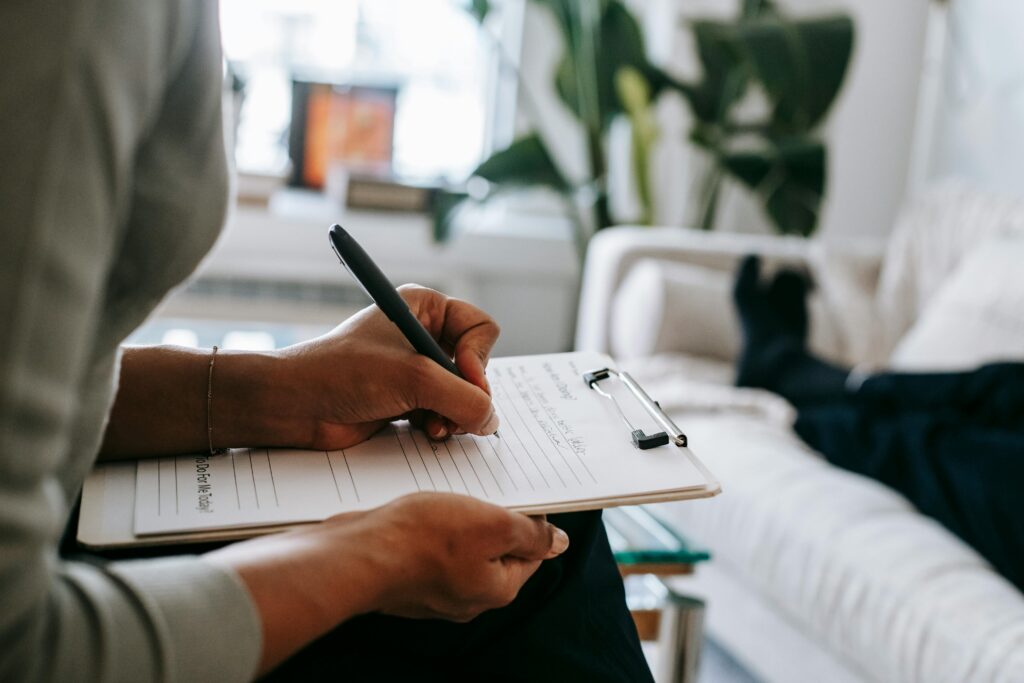 pexels-photo-5699456-5699456 Unrecognizable ethnic female therapist taking notes on clipboard while filling out form during psychological appointment with anonymous client lying on blurred background